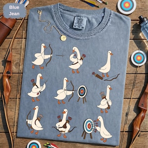 Peut inclure: Un t-shirt bleu jean avec une impression graphique d'oies blanches dans diverses poses de tir à l'arc. Les oies sont représentées tenant des arcs, portant des carquois de flèches et visant des cibles circulaires. Le t-shirt a un col rond.