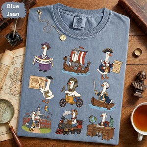 Peut inclure: Un t-shirt bleu jean avec une impression graphique représentant neuf oies historiques. Les illustrations montrent des oies déguisées en suffragette, viking, personnage colonial, érudit, pharaon égyptien, soldat, fondateur, conducteur de train et homme d'État à un bureau.