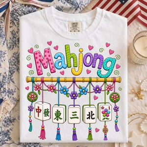 Peut inclure: Un t-shirt blanc avec le mot Mahjong écrit en lettres colorées et à motifs au-dessus d'un graphique représentant une tige de bambou. Cinq tuiles de mah-jong avec des caractères chinois et des motifs floraux pendent, ornées de pompons et cœurs.