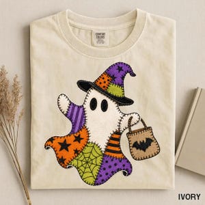 Peut inclure: Un t-shirt ivoire avec un motif central représentant un fantôme portant un chapeau de sorcière en patchwork. Le fantôme est composé de sections de tissu à motifs orange, violet et vert, et porte un petit sac beige avec une chauve-souris noire.