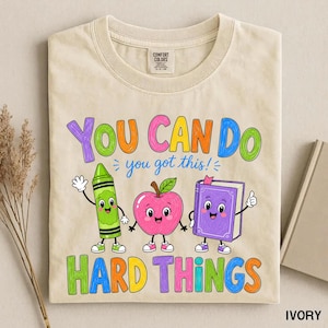 Peut inclure: Un t-shirt ivoire avec un motif coloré portant le texte "YOU CAN DO hard things" et "you got this!". Le design comprend un crayon vert, une pomme rose et un livre violet, tous dotés de visages souriants, de bras et de jambes de style dessin animé.