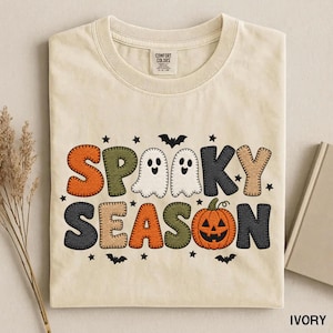 Peut inclure: Un t-shirt ivoire arborant un motif avec le texte "SPOOKY SEASON" dans une police style point de couture. Le design comprend deux fantômes blancs, une citrouille orange vif, ainsi que de petites chauves-souris et étoiles noires.