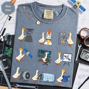 Peut inclure: Un t-shirt bleu jean avec une impression graphique de neuf oies de dessin animé effectuant diverses tâches d'ingénierie aérospatiale, travaillant sur des moteurs de fusée, des satellites et des panneaux de contrôle, avec une étiquette Comfort Colors XL.