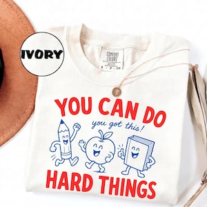Peut inclure: Un t-shirt ivoire avec un motif représentant un crayon, une pomme et un livre souriants. Le texte "YOU CAN DO HARD THINGS" est écrit en rouge et "you got this!" en bleu. Le t-shirt porte une étiquette Comfort Colors et une étiquette ronde.