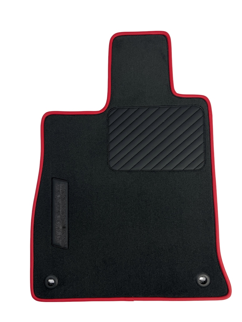 May include: Black car floor mat with a red trim. The mat has a quilted black section and two metal grommets. The mat is designed to fit the driver's side of a vehicle.