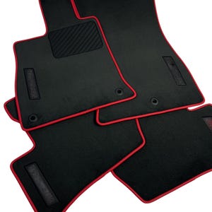 May include: Set of four black car floor mats with a red trim. The mats have a textured, non-slip surface and are designed to fit the contours of a car's floor. The mats are made of a durable material and are designed to protect the car's interior.