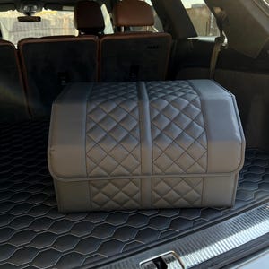 May include: A black, quilted car trunk organizer sits in the trunk of a vehicle. The organizer has a diamond pattern and is made of a leather-like material. The trunk also features a black mat with a white stitched hexagonal pattern.