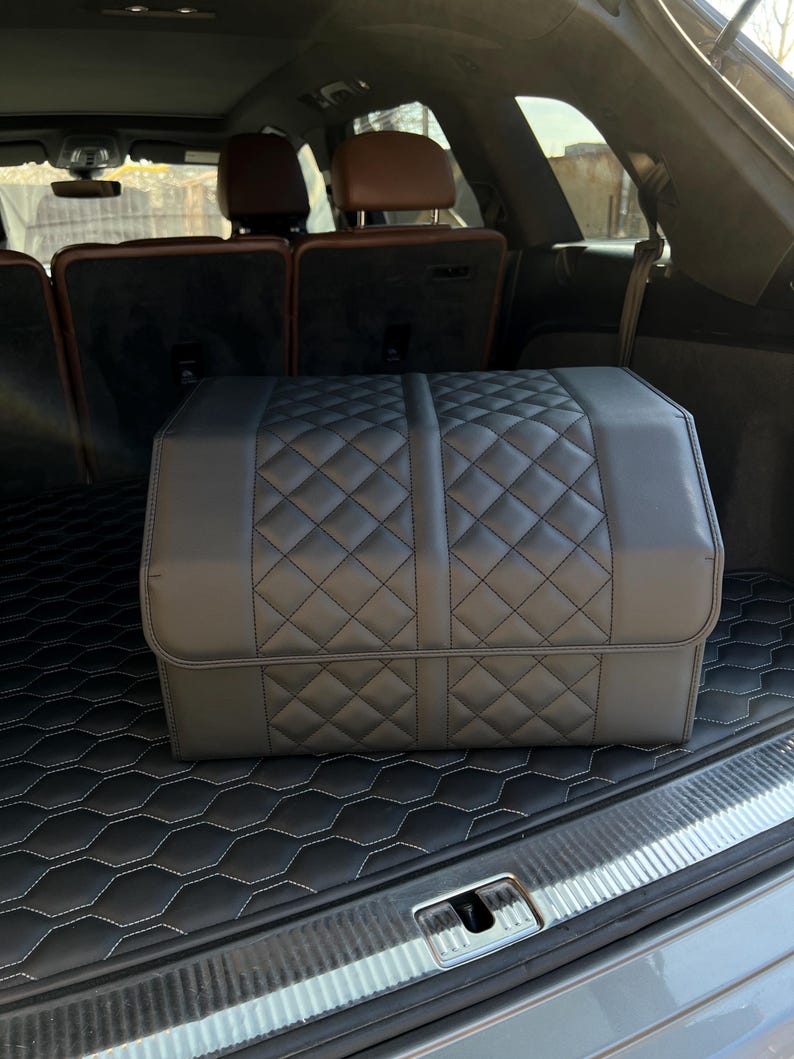 May include: A black, quilted car trunk organizer sits in the trunk of a vehicle. The organizer has a diamond pattern and is made of a leather-like material. The trunk also features a black mat with a white stitched hexagonal pattern.