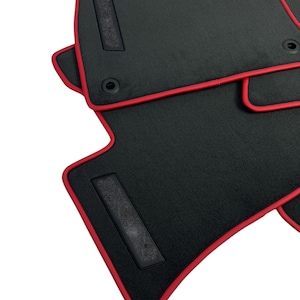 May include: Black car floor mats with a red trim. The mats have a textured surface and a rectangular patch with a grid pattern. The mats are designed to fit the contours of a car's floor.