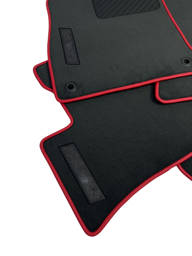 May include: Black car floor mats with a red trim. The mats have a textured surface and a rectangular patch with a grid pattern. The mats are designed to fit the contours of a car's floor.