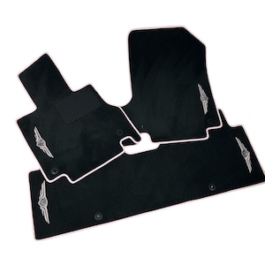 May include: Four black car floor mats with white edging. Each mat has a silver emblem. The mats are designed for the front and rear footwells of a vehicle. The mats are made of a durable material.