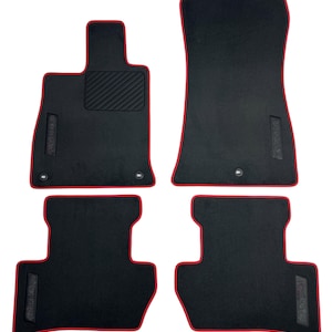 May include: Set of four black car floor mats with red trim. The mats have a textured surface and are designed to fit a vehicle's interior. The set includes two front and two rear mats.