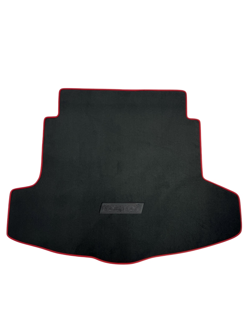 May include: Black car trunk mat with a red trim. The mat has a rectangular shape with a contoured design to fit the trunk. A rectangular textured patch is centered on the mat.