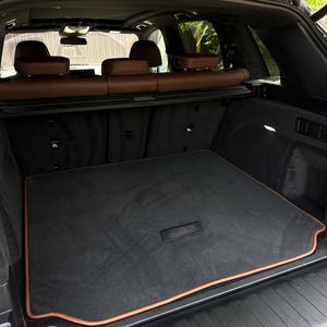 May include: A black car trunk with a black carpeted floor and a brown trim. The car's interior features brown leather seats and a panoramic sunroof. The trunk is open, revealing the cargo area.