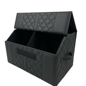 May include: Black, quilted car organizer with two compartments and a lid. The organizer has a rectangular shape with a diamond pattern and a handle on the side. A velcro strap is on the front.