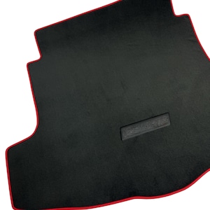 May include: Black car floor mat with a red trim. The mat has a rectangular shape with a custom fit design. The mat has a textured surface and a small rectangular patch in the center.