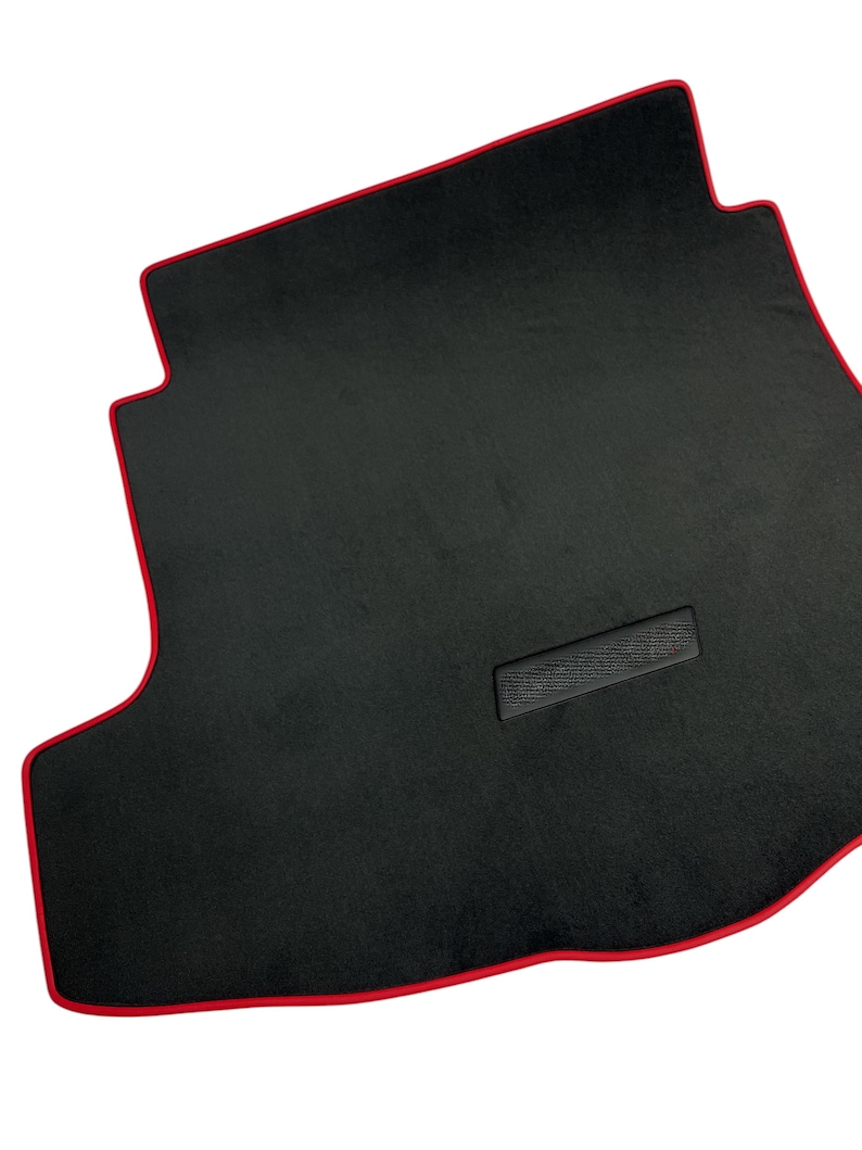 May include: Black car floor mat with a red trim. The mat has a rectangular shape with a custom fit design. The mat has a textured surface and a small rectangular patch in the center.