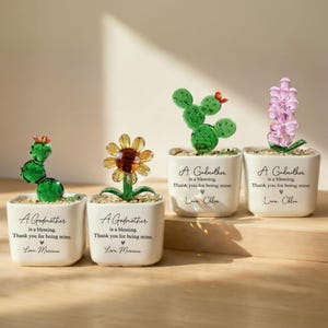 May include: Four small, white ceramic pots with glass plant sculptures. The plants include a cactus, a sunflower, a cactus, and a lavender-like flower. Each pot has the text "A Godmother is a blessing. Thank you for being mine. Love, [Name]" printed on it.