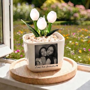 Może przedstawiać: Biała ceramiczna doniczka ze zdjęciem dw&oacute;ch kobiet, trzymających szklaną kompozycję kwiatową z tulipan&oacute;w. Na doniczce widnieje napis "Long live friendship". Kompozycja zawiera trzy białe kwiaty tulipan&oacute;w i zielone liście.