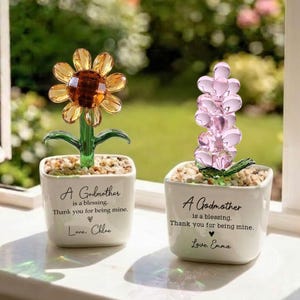 May include: Two decorative glass flower sculptures in white square pots. One is a yellow sunflower, the other is a pink flower. Each pot has the text "A Godmother is a blessing. Thank you for being mine. Love, Chloe/Emma."