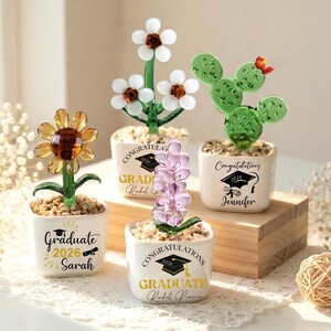 May include: A collection of decorative glass flower and cactus sculptures in small white pots. The sunflower, daisy, and lavender-like sculptures are in full bloom. Each pot has a graduation-themed message with a name and the year 2026.
