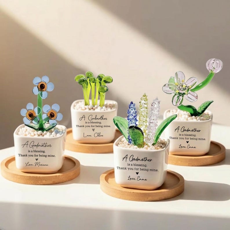 May include: Four decorative glass flower arrangements in white square pots on wooden coasters. Each pot has the text "A Godmother is a blessing. Thank you for being mine." and a name. The flowers are blue, green, and clear.