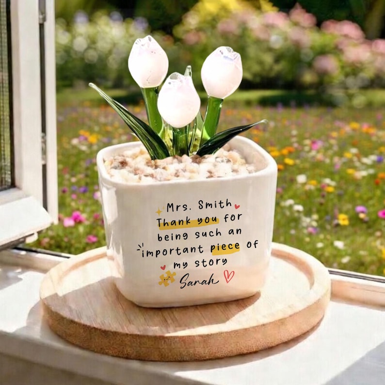 Puede incluir: Una maceta de cer&aacute;mica blanca con un arreglo de tulipanes de cristal. La maceta tiene el texto "Sra. Smith, Gracias por ser una parte tan importante de mi historia, Sarah". La maceta est&aacute; sobre una bandeja de madera.