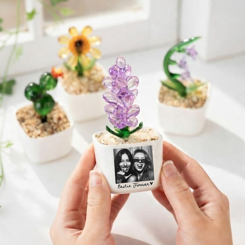 Może przedstawiać: Mała biała ceramiczna doniczka ze zdjęciem dw&oacute;ch kobiet i napisem "Bestie Forever V". Doniczka zawiera fioletowy kryształowy kwiat. Inne doniczki ze szklanymi kwiatami są w tle.