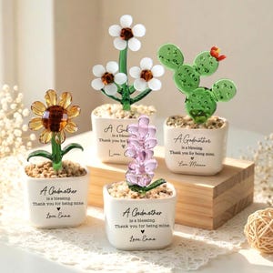 May include: A collection of glass flower sculptures in small white pots. The flowers include a sunflower, daisies, a cactus, and lavender. Each pot has the text "A Godmother is a blessing. Thank you for being mine."