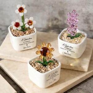 May include: Three small, white ceramic pots each hold a different glass flower arrangement. One has white flowers with red centers, another has a lavender arrangement, and the third has a yellow sunflower. Each pot has text that reads "A Godmother is a blessing. Thank you for being mine."