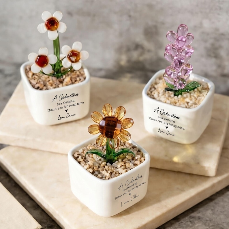 May include: Three small, white ceramic pots each hold a different glass flower arrangement. One has white flowers with red centers, another has a lavender arrangement, and the third has a yellow sunflower. Each pot has text that reads "A Godmother is a blessing. Thank you for being mine."
