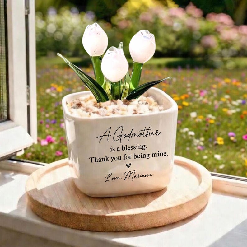 May include: A white ceramic pot with a floral arrangement of white tulip-shaped glass flowers. The pot has the text "A Godmother is a blessing. Thank you for being mine. Love, Mariana."