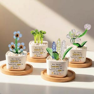 Puede incluir: Cuatro arreglos florales decorativos de vidrio en peque&ntilde;as macetas cuadradas blancas sobre posavasos de madera. Cada maceta tiene un mensaje escrito a mano. Las flores son de varios colores y estilos, incluyendo vidrio azul, verde y transparente.