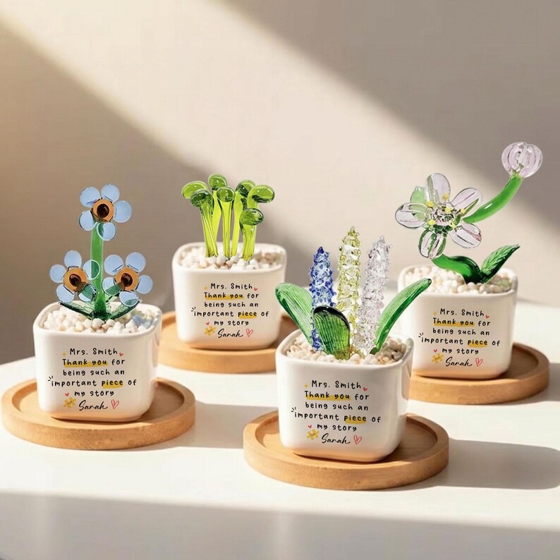 Puede incluir: Cuatro arreglos florales decorativos de vidrio en peque&ntilde;as macetas cuadradas blancas sobre posavasos de madera. Cada maceta tiene un mensaje escrito a mano. Las flores son de varios colores y estilos, incluyendo vidrio azul, verde y transparente.