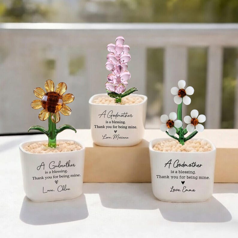 May include: Three decorative glass flower arrangements in white square pots. The flowers include a sunflower, a pink flower, and white flowers. Each pot has the text "A Godmother is a blessing. Thank you for being mine." and a name.