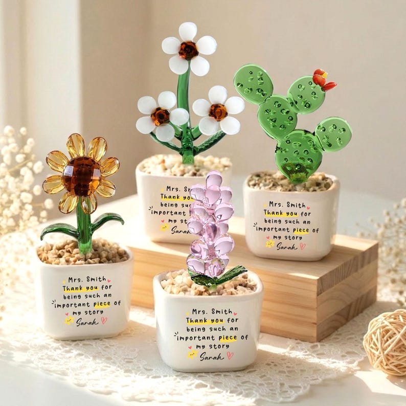 Puede incluir: Una colecci&oacute;n de esculturas de flores de vidrio en macetas blancas peque&ntilde;as. Las flores incluyen un girasol, margaritas, una planta parecida a la lavanda y un cactus. Cada maceta tiene el texto "Mrs. Smith, Thank you for being such an important piece of my story, Sarah."