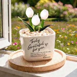 Peut inclure: Un pot en céramique blanc avec un arrangement floral de trois tulipes en verre blanc. Le pot porte l'inscription "Truly Grateful for you." et un message. L'arrangement repose sur un plateau en bois, avec un arrière-plan de jardin flou.