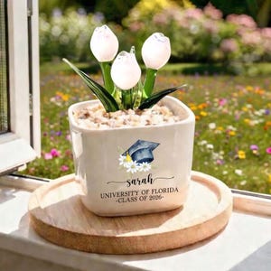 May include: An off-white ceramic pot with a graduation cap and floral design, containing glass tulip flowers. The pot is inscribed with "Sarah", "University of Florida", and "Class of 2026". It rests on a round wooden tray, with a blurred floral background.