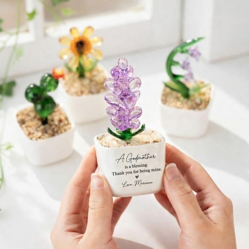 May include: A collection of miniature glass flower sculptures in white ceramic pots. The central pot features a purple flower and the text "A Godmother is a blessing. Thank you for being mine. Love, Mariana."