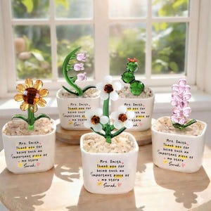 Puede incluir: Cinco esculturas de flores de cristal en macetas blancas. Las flores incluyen un girasol, margaritas, un cactus y otros dise&ntilde;os florales. Cada maceta tiene el texto "Sra. Smith, Gracias por ser una parte tan importante de mi historia, Sarah."