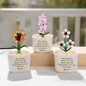 Puede incluir: Tres esculturas de flores de cristal en macetas blancas. Una es un girasol, otra una flor parecida a la lavanda y la tercera tiene flores blancas. Cada maceta tiene el texto "Sra. Smith, Gracias por ser una parte importante de mi historia, Sarah."