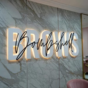 Könnte beinhalten: Ein Schild mit der Aufschrift "Brows Bombshell" in einem Schönheitssalon. Das Wort "Brows" ist in großen, weißen Blockbuchstaben mit einem warmen, gelben Schein dahinter geschrieben. Das Wort "Bombshell" ist in schwarzer Schreibschrift.
