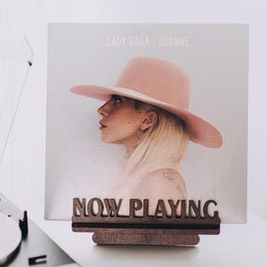 May include: A record player with a white vinyl record is next to a wooden "NOW PLAYING" sign holding a Lady Gaga "Joanne" album cover. The album cover features a woman wearing a pink hat.