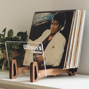 Può includere: Un portadischi in legno e acrilico che mostra una collezione di vinili. Il supporto presenta il testo "Willson's Collection" e "Best Husband Ever". La copertina dell'album "Thriller" di Michael Jackson è visibile.