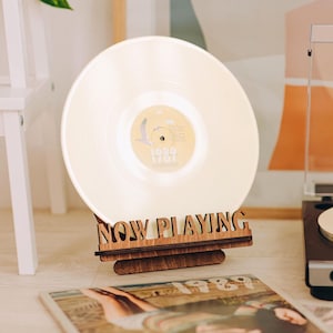 May include: A dark brown wooden display stand featuring the laser-cut text "NOW PLAYING" in a bold font. The stand holds a cream-colored vinyl record upright, showcasing a central label with a bird illustration and the text "1989".
