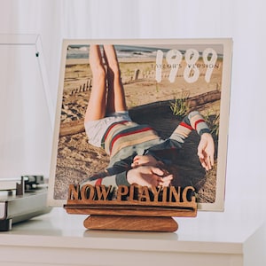 May include: A wooden record stand displaying a vinyl record with the text "1989 Taylor's Version" and "NOW PLAYING". The record cover features a person lying on a beach. The stand is on a white surface.