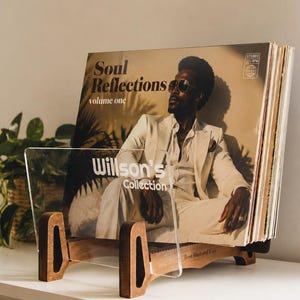 Op de afbeelding: Een houten vinylplatenstandaard met een doorzichtig acryl voorpaneel met de witte tekst Willson's Collection. De standaard bevat een stapel vinylplaten, waaronder een hoes met de titel Soul Reflections volume one met een persoon in een pak.