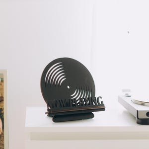 May include: A black wooden display stand featuring a circular vinyl record cutout design with concentric grooves. The base of the stand includes the text NOW PLAYING in a bold, cut-out font, designed to hold a record sleeve in an upright position.