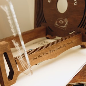 May include: A dark brown wooden vinyl record storage rack featuring a laser-engraved inscription that reads For the One Who Rocks My World. The rack includes a circular cutout and a graphic of a record player on the back panel with grooved slots.