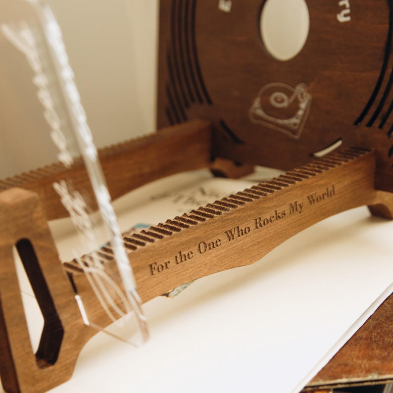 May include: A dark brown wooden vinyl record storage rack featuring a laser-engraved inscription that reads For the One Who Rocks My World. The rack includes a circular cutout and a graphic of a record player on the back panel with grooved slots.